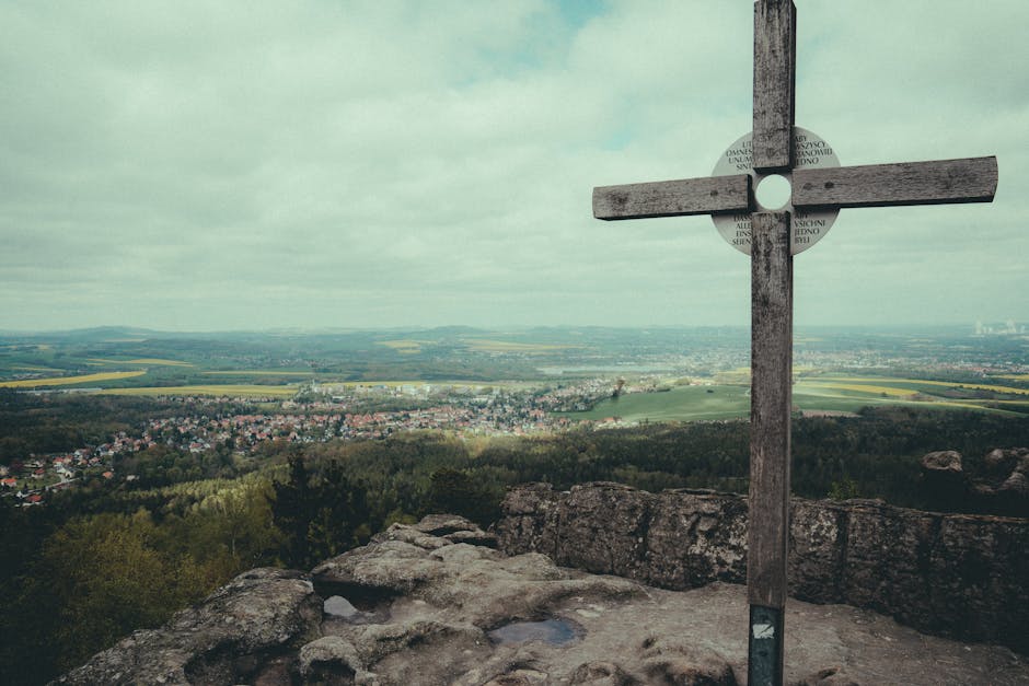 A serene view of a wooden cross standing on a rocky outcrop with Zittau valley in the background.